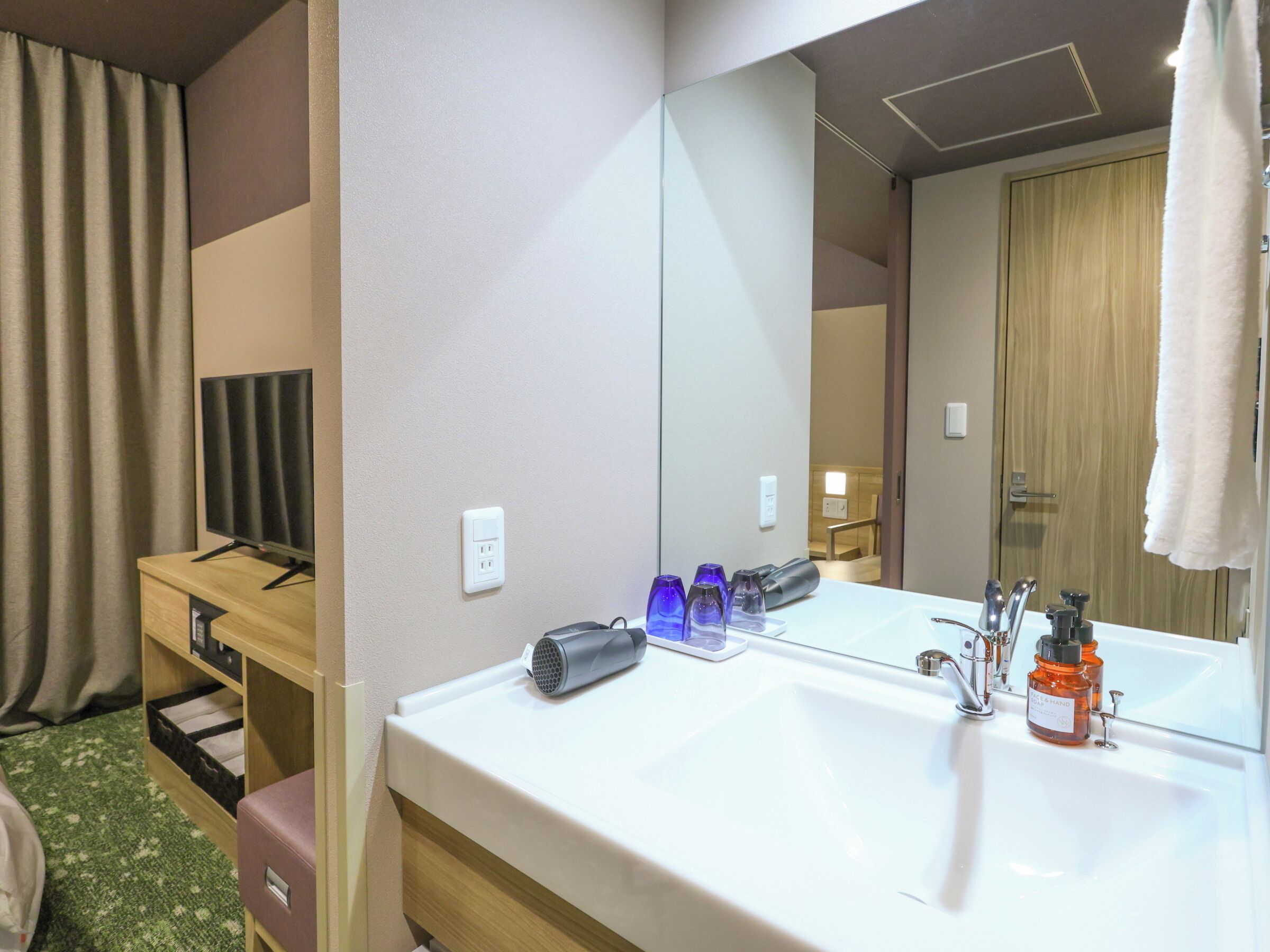 A hotel bathroom with a white sink, a mirror, a towel hanging on the right, and a television on a wooden cabinet.