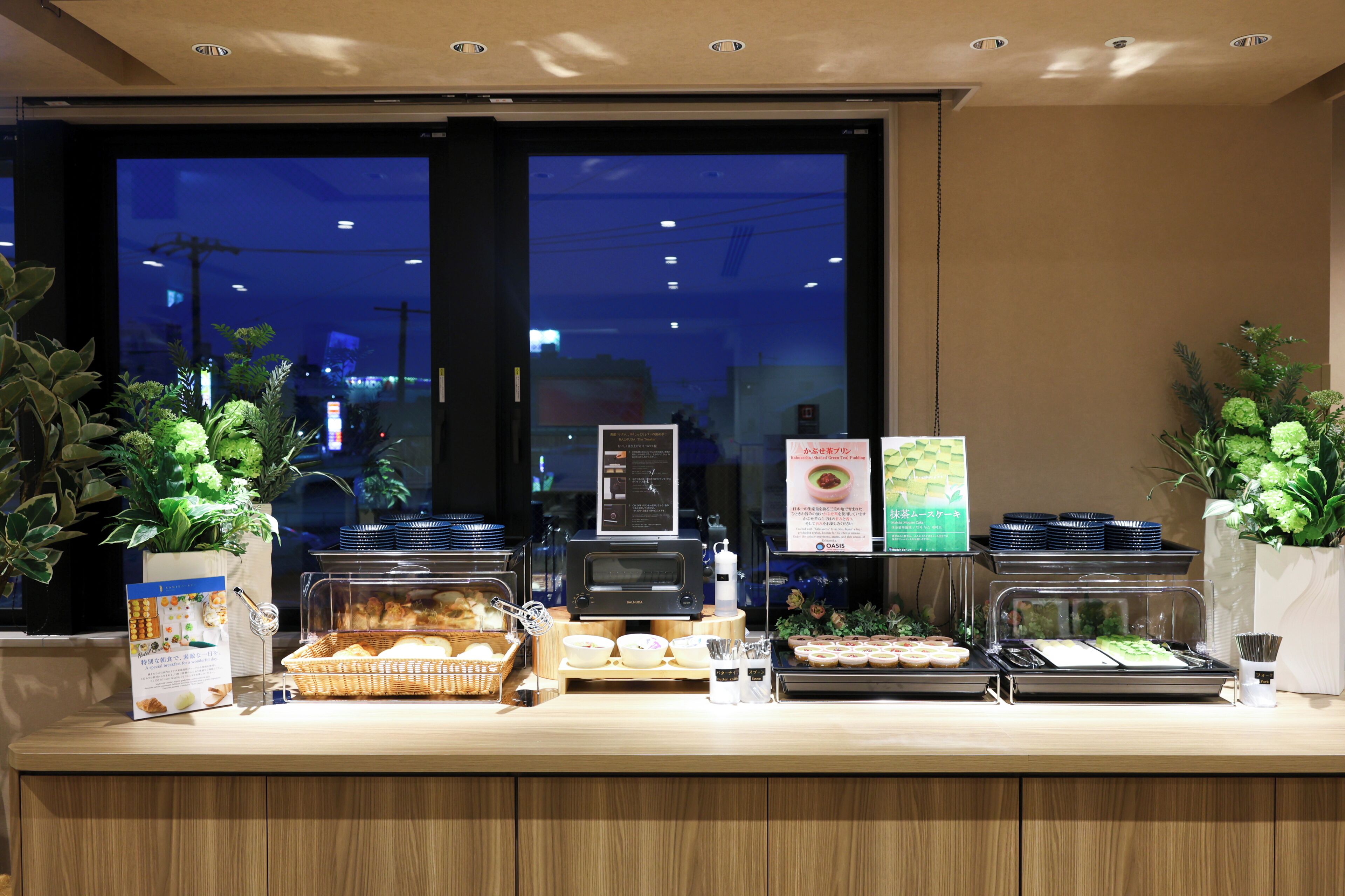 Mostrador de buffet con diferentes alimentos, un panel de menú digital y plantas en macetas.