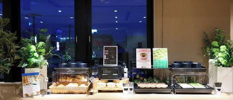 Mostrador de buffet con diferentes alimentos, un panel de menú digital y plantas en macetas.