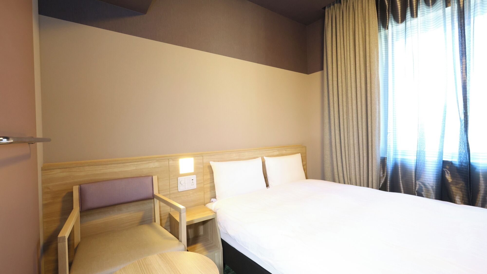 Una habitación de hotel con una cama, una silla, una mesita de noche y un ventanal con cortinas.