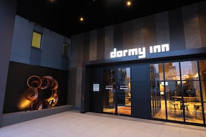 Un edificio moderno con un letrero de 'Dormy Inn', grandes ventanales de vidrio y una escultura metálica a la izquierda.