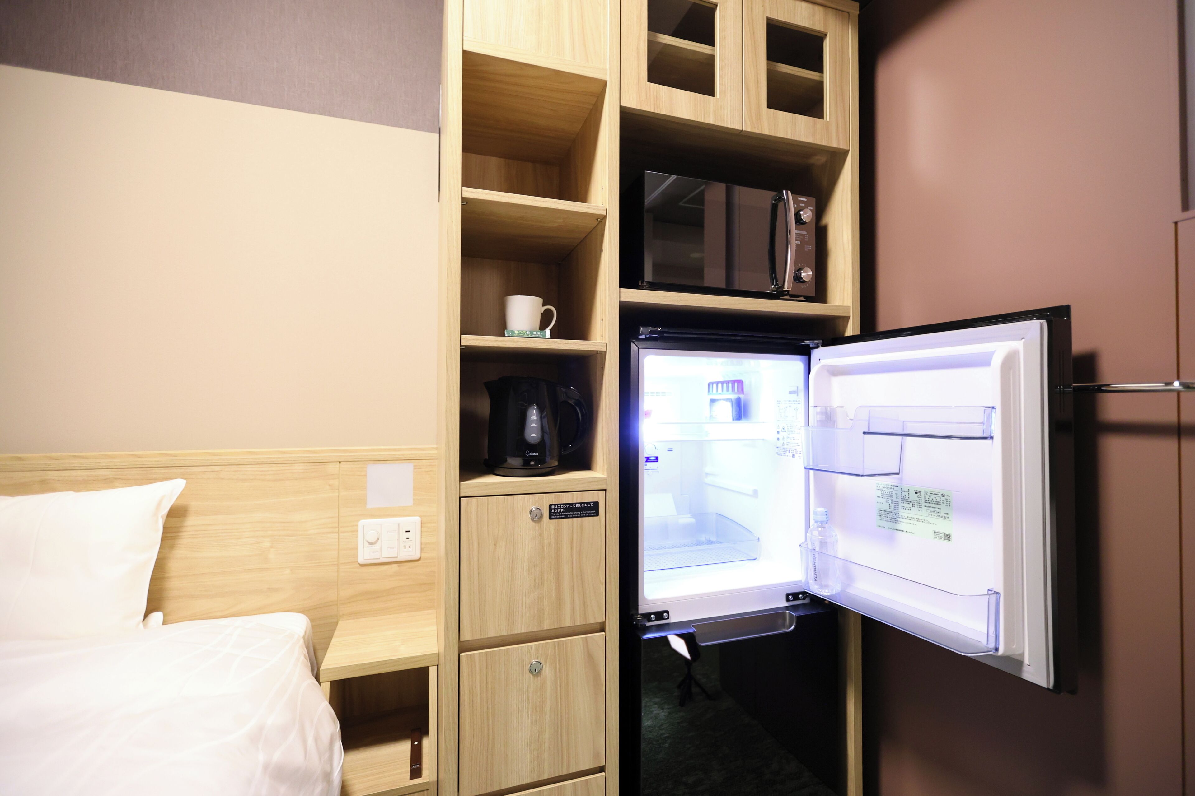 Habitación de hotel con cama, un mueble de madera con cafetera y un refrigerador con pantalla digital.