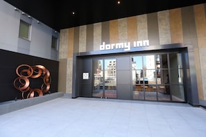 Ein modernes Gebäude mit einem 'Dormy Inn' Schild, großen Glasüren und einer Metallskulptur auf der linken Seite.