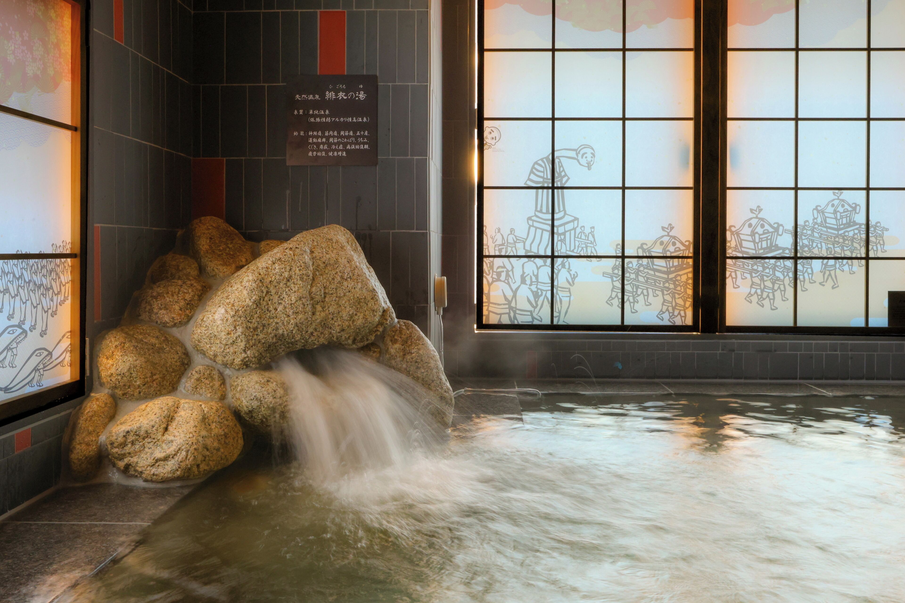A traditional Japanese bath with large rocks and flowing water, a tiled wall with calligraphy, and a window with a drawing of people in historical attire.