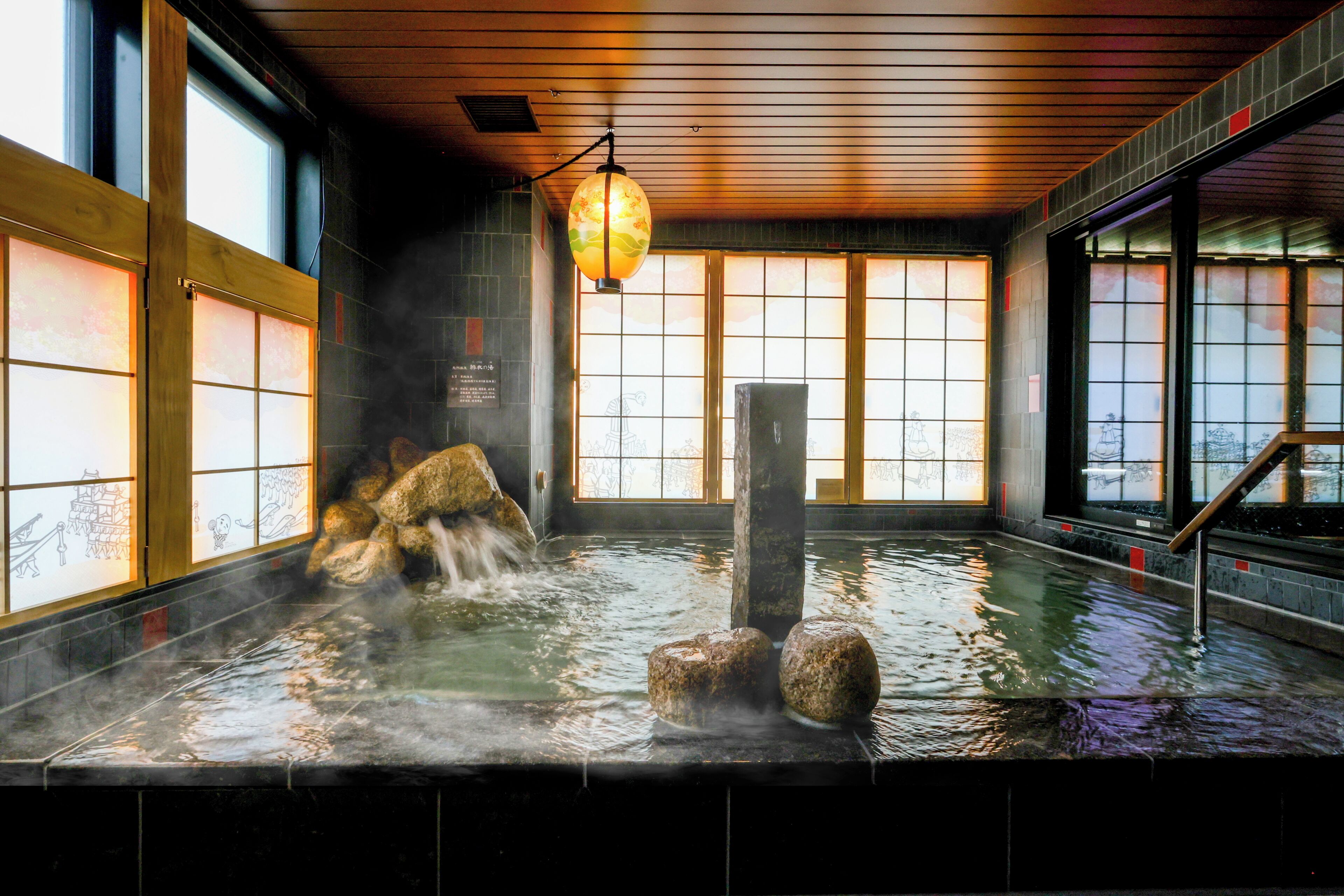 A traditional Japanese onsen with a wooden interior, a stone hot spring, and a view of the city through large windows.