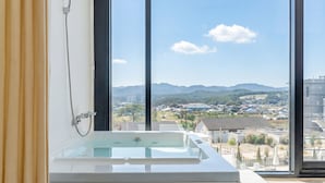 Un baño moderno con bañera acristalada, ventanas amplias y vistas a un paisaje urbano y montañas.