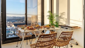 Un comedor con una mesa dispuesta para una comida, sillas de mimbre, una planta en maceta y vistas de la ciudad a través de amplios ventanales.