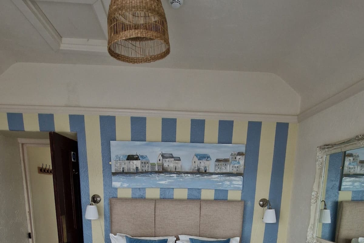 A hotel room with a bed, a headboard with a painting, a hanging light fixture, and a mirror on the wall.