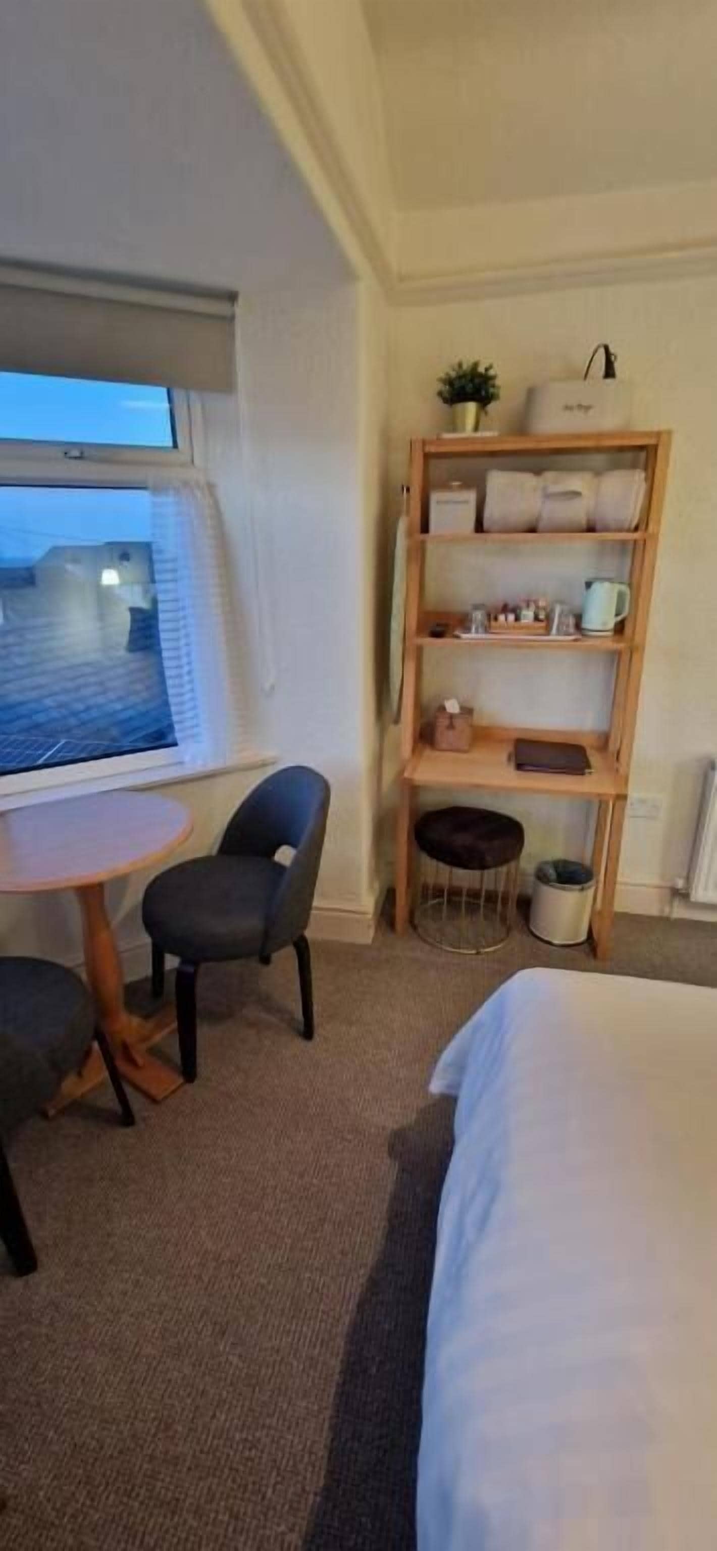 Ein Hotelzimmer mit einem Bett, einem kleinen runden Tisch mit zwei Stühlen, einer Holzablage mit diversen Gegenständen, einem Fenster mit Jalousien und Blick nach draußen.