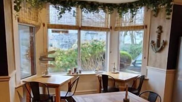 A dining area with a wooden table and chairs, large windows with curtains, and a ceiling fan. - Black Sheep Lodge (Holyhead)