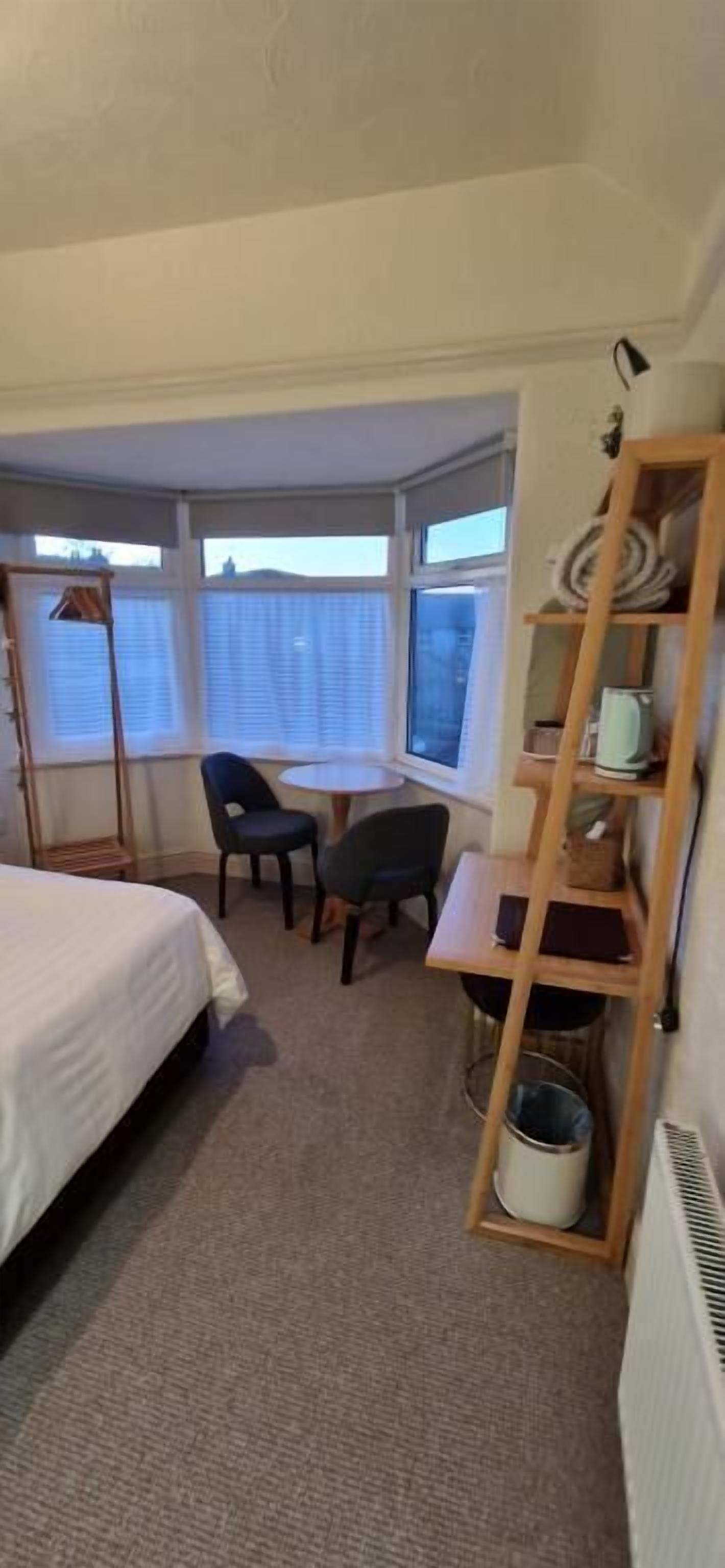 A hotel room with a bed, a desk, two chairs, a small table, a shelf with items, and a window with blinds.