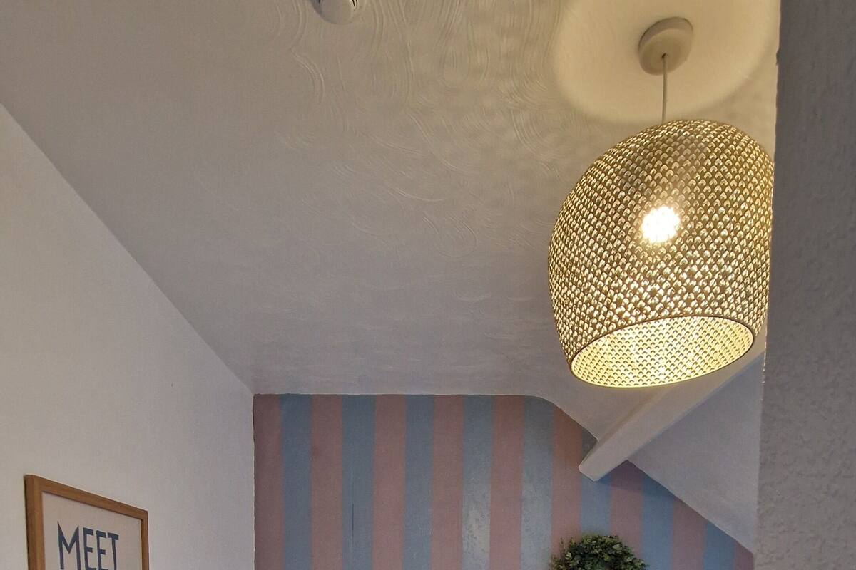 A bedroom with a bed, a nightstand, a wall-mounted lamp, a 'Meet Me At The Beach' sign, and a striped wall.