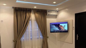 Una habitació d'hotel moderna amb un llit gran, cortines beix i un televisor fixat a la paret, així com un aparell d'aire condicionat.
