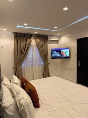 A modern hotel room with a large bed, beige curtains, a mounted TV, and an air conditioning unit on the wall. - 4-bedroom house in enchanting Victoria Island with AC (Victoria Island)