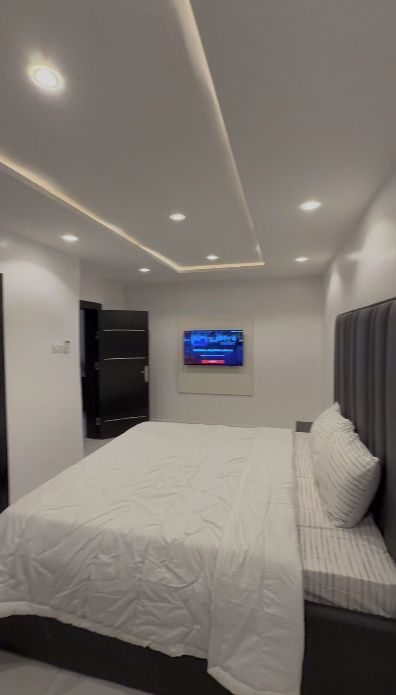 Une chambre à coucher moderne, avec un lit, un téléviseur fixé au mur et un éclairage encastré au plafond.