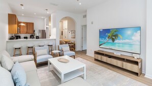 Ein modernes Wohnzimmer mit großem Flachbildfernseher, einer Couch, einem Couchtisch und einer Küchenzeile mit Holzschränken und einer Essgruppe.