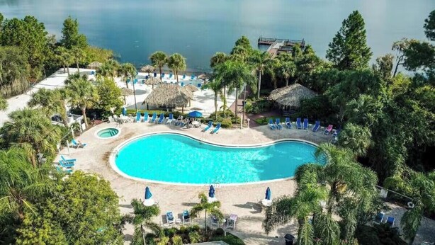 Ein Resort mit einem großen Schwimmbecken, umgeben von Liegestühlen und Sonnenschirmen, vor einer Kulisse aus ruhigem See und dichtem Baumbestand.