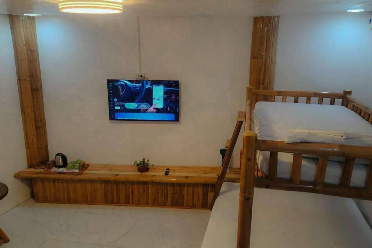 A room with a bunk bed, a wooden bench with a kettle and a plant, and a flat-screen TV mounted on the wall.