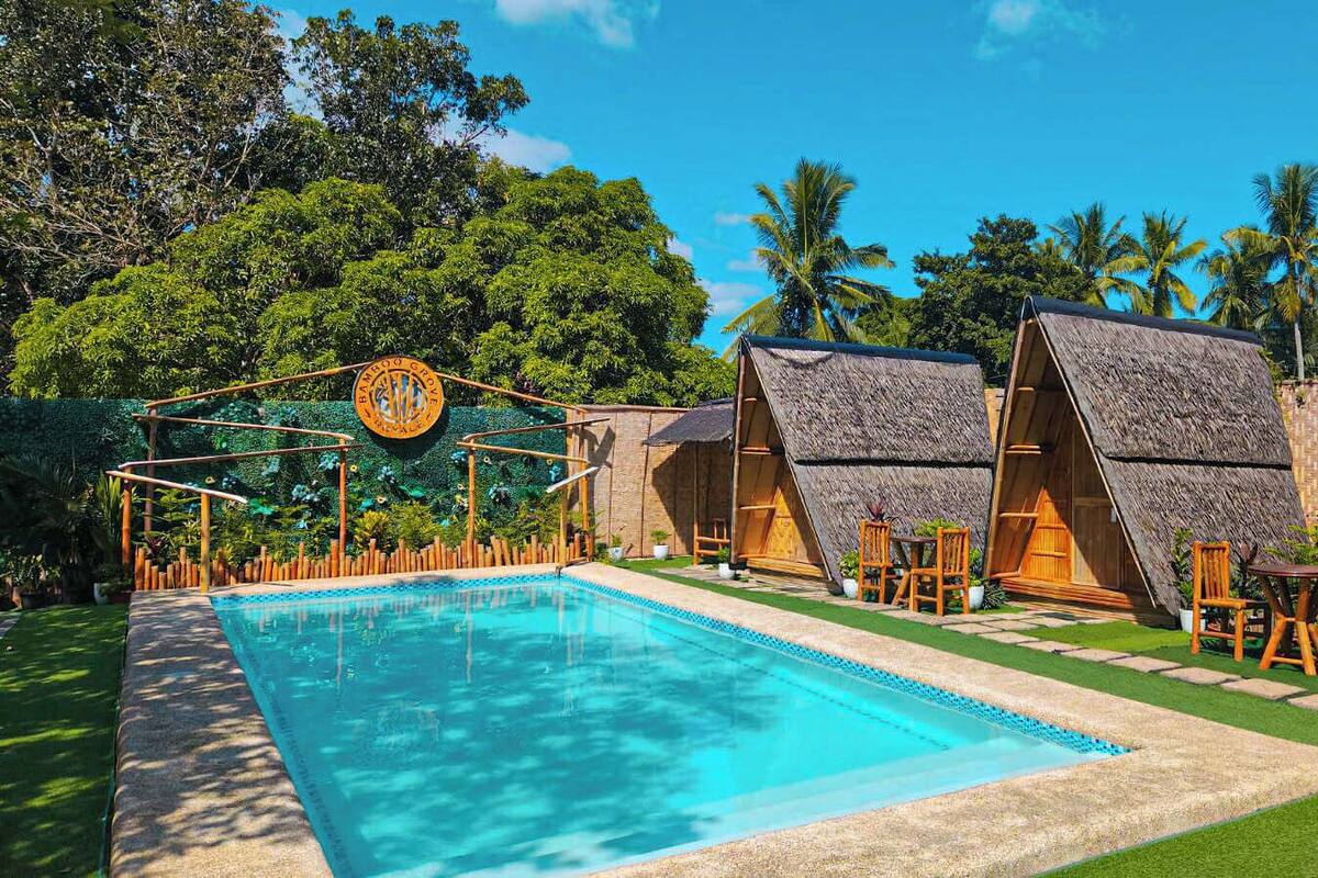 A pool area with thatched-roof huts, a wooden fence, and lush greenery.