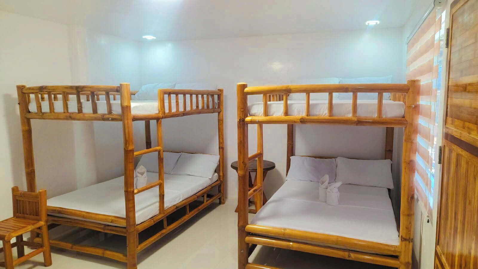 Dos literas con estructura de madera y ropa de cama blanca, una silla de madera pequeña y una mesa redonda en una habitación con paredes blancas y una puerta de madera.