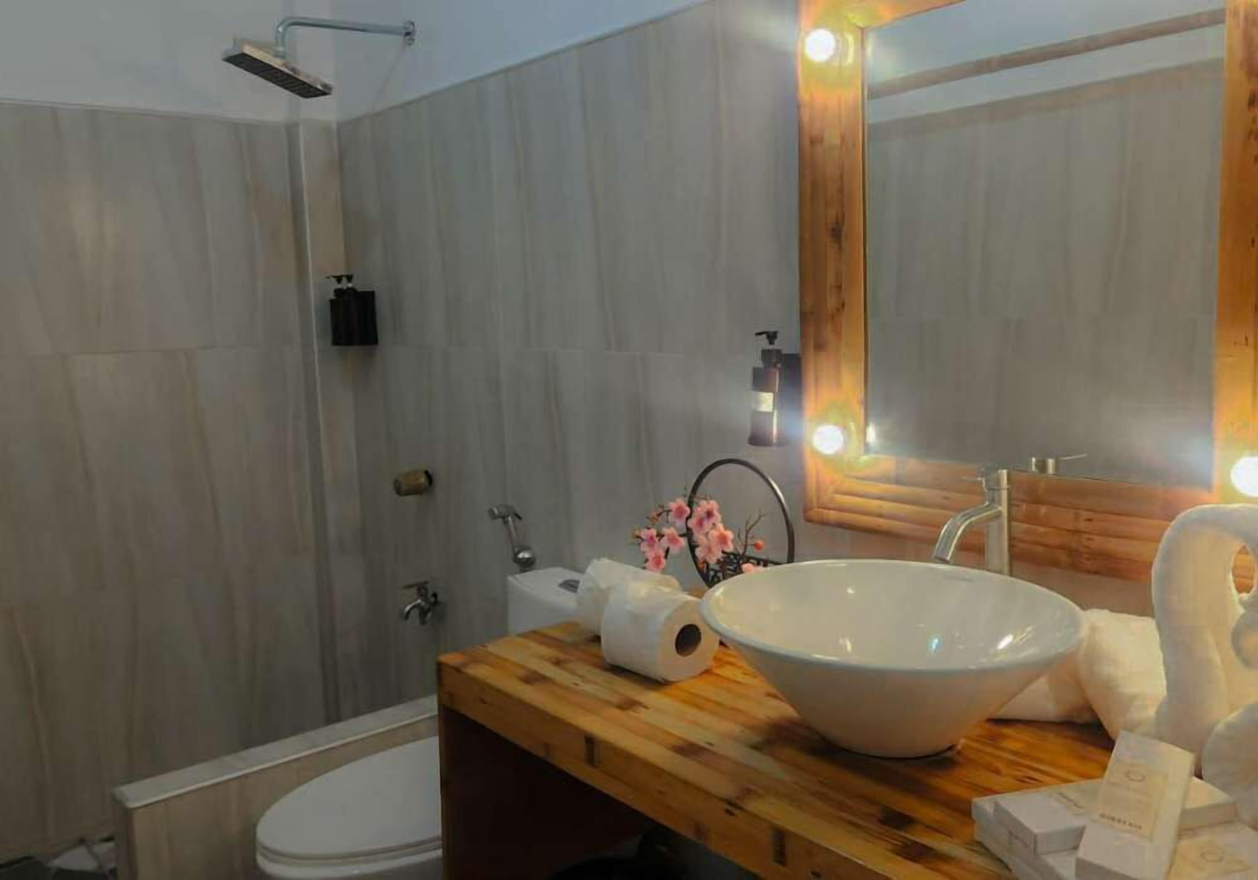 Un baño moderno con tocador de madera, un lavamanos blanco grande, un espejo con luces, una ducha y un inodoro.