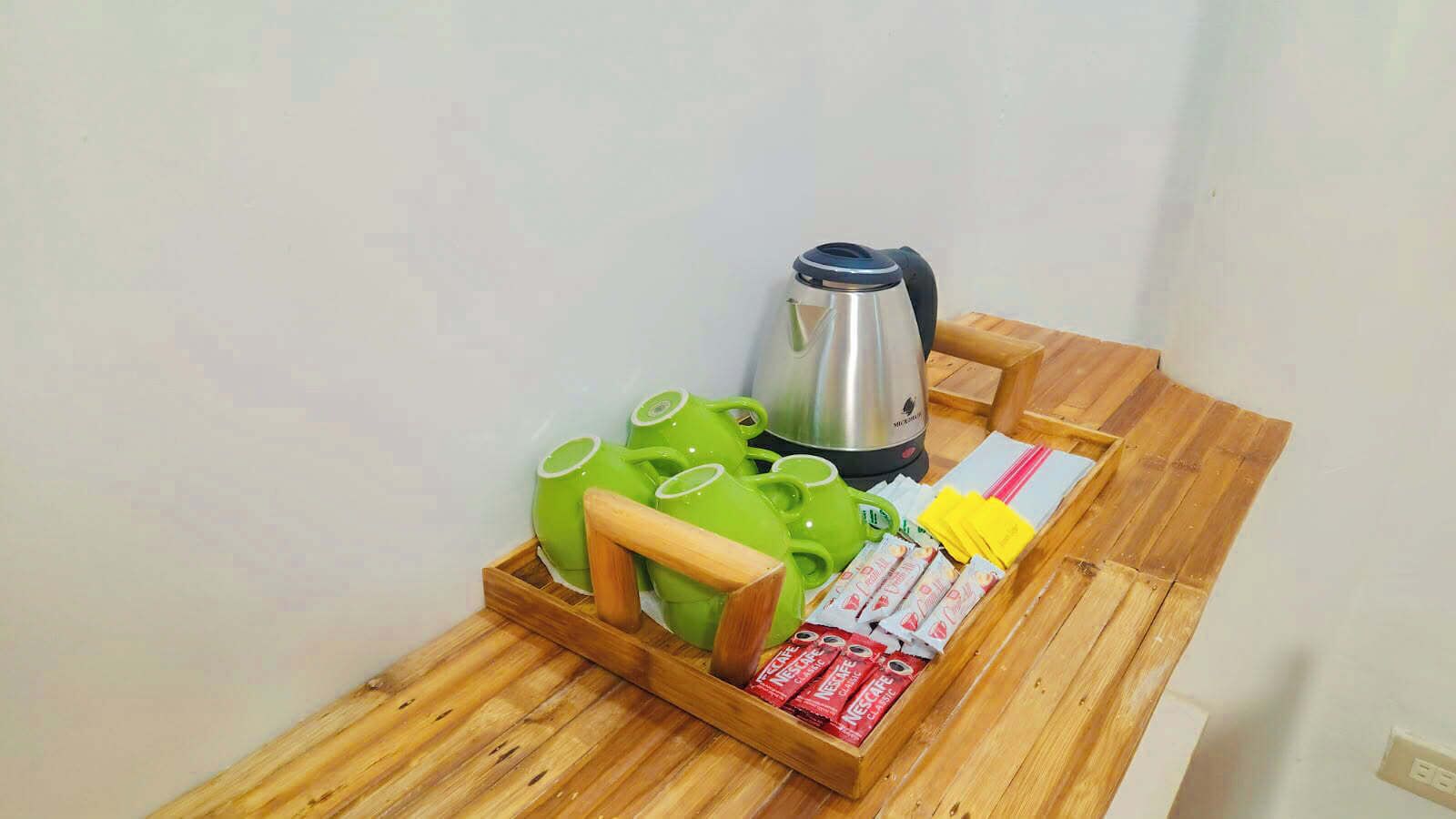 Una mesa de madera con un hervidor de acero inoxidable, teteras verdes, una bandeja de madera y sobres de café instantáneo.