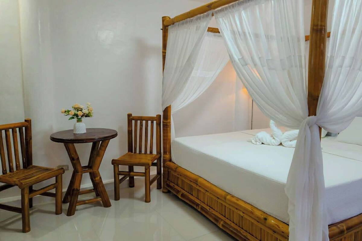 A room with a four-poster bed, two wooden chairs, a small round table, and a vase with flowers.