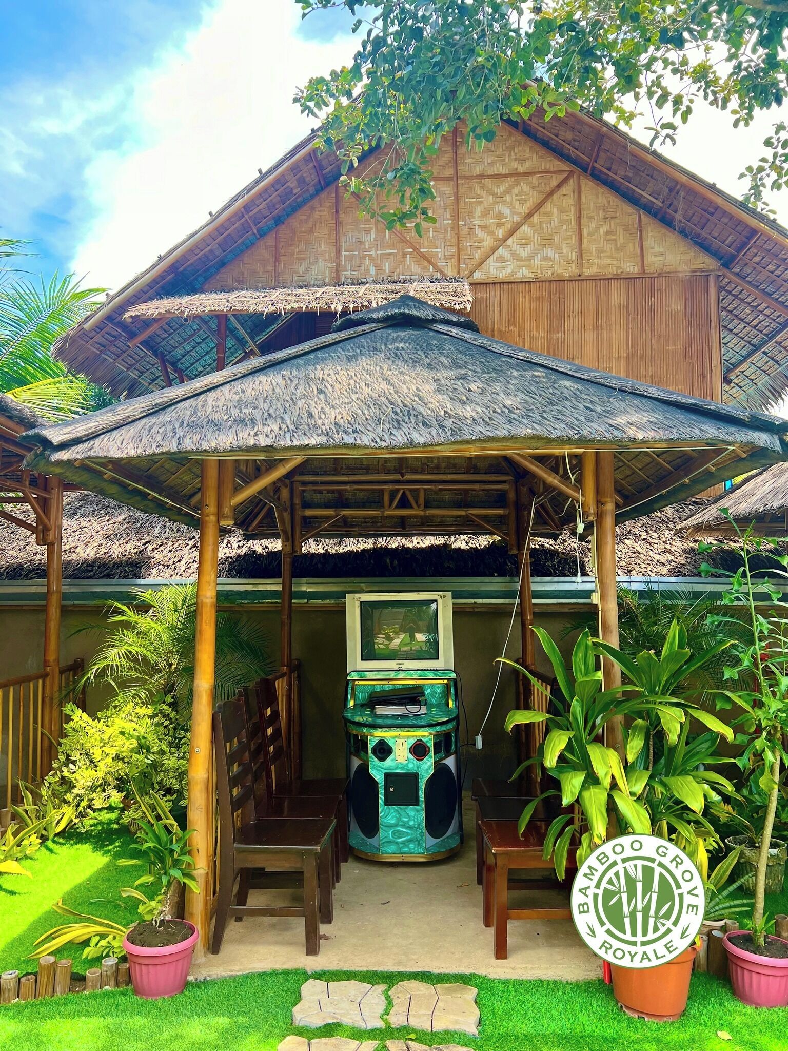 A covered outdoor seating area with a thatched roof, surrounded by potted plants and a wooden structure.