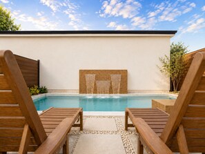 A pool area with wooden lounge chairs, a clear blue pool, and a wooden wall with a water feature. - Burbank Getaway (Burbank)