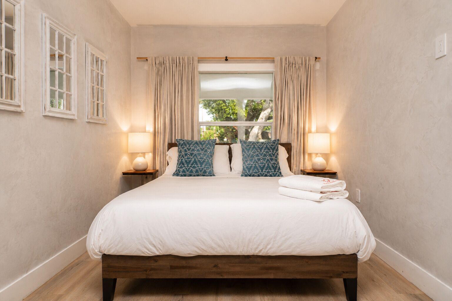 Un dormitorio con cama de madera, dos lámparas de noche, una ventana con cortinas y vista a la vegetación.