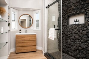 A bathroom with a wooden vanity, a round mirror, and a glass shower enclosure with a stone wall. - Burbank Getaway (Burbank)