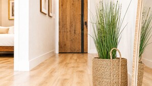 A wicker basket with green plants on a wooden floor, a wooden door, and a white wall with framed pictures.