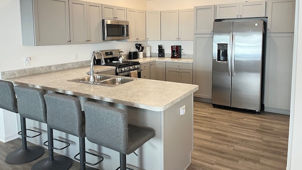 Una cocina moderna con armarios grises, electrodomésticos de acero inoxidable, una isla central con taburetes y suelo de madera.