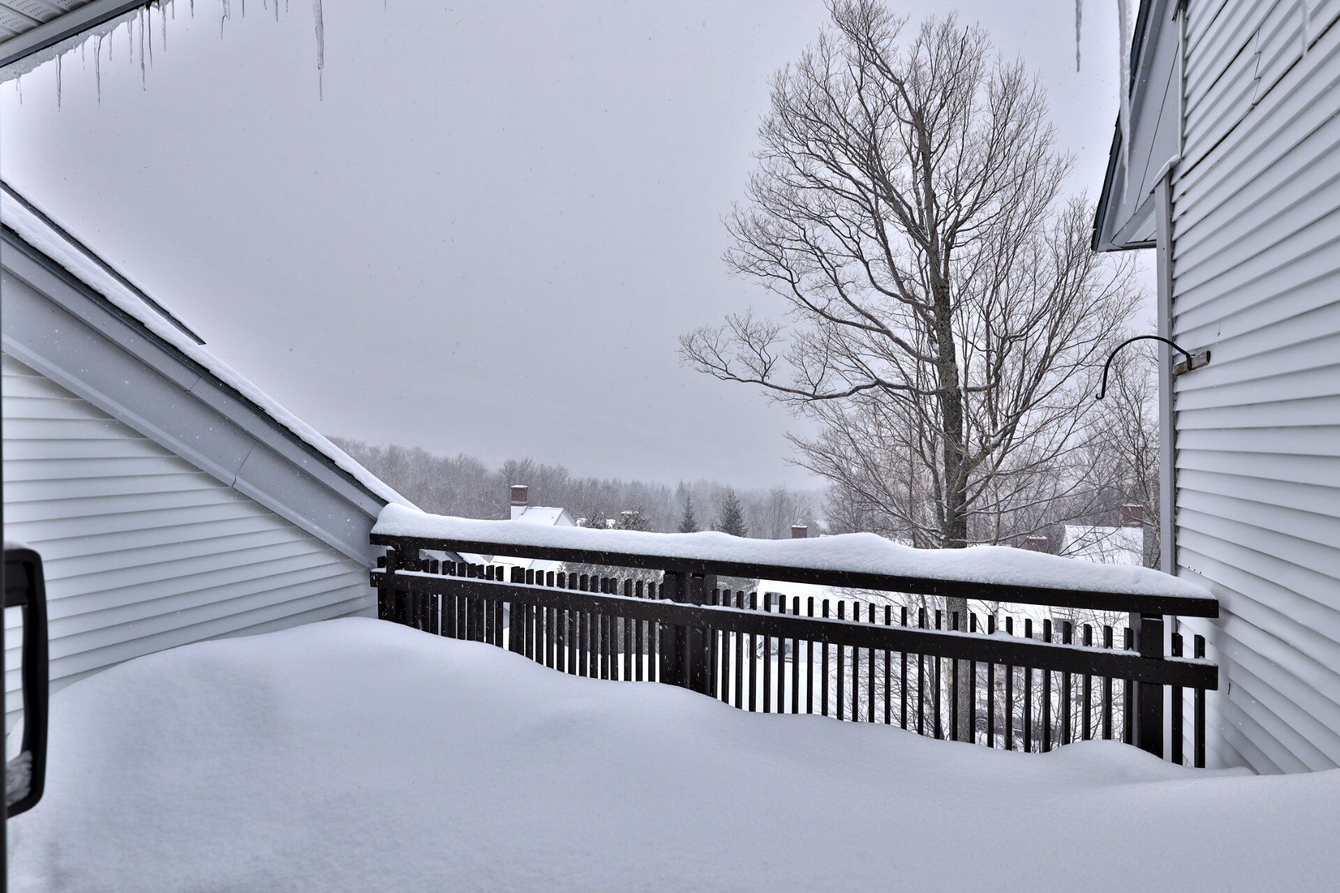 Un balcón cubierto de nieve con baranda de madera, un árbol sin hojas y una casa al fondo.