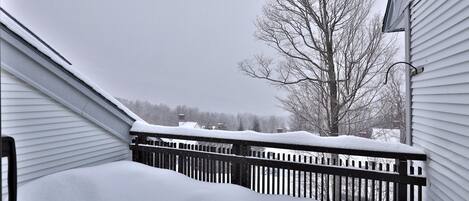 Un balcón cubierto de nieve con baranda de madera, un árbol sin hojas y una casa al fondo.