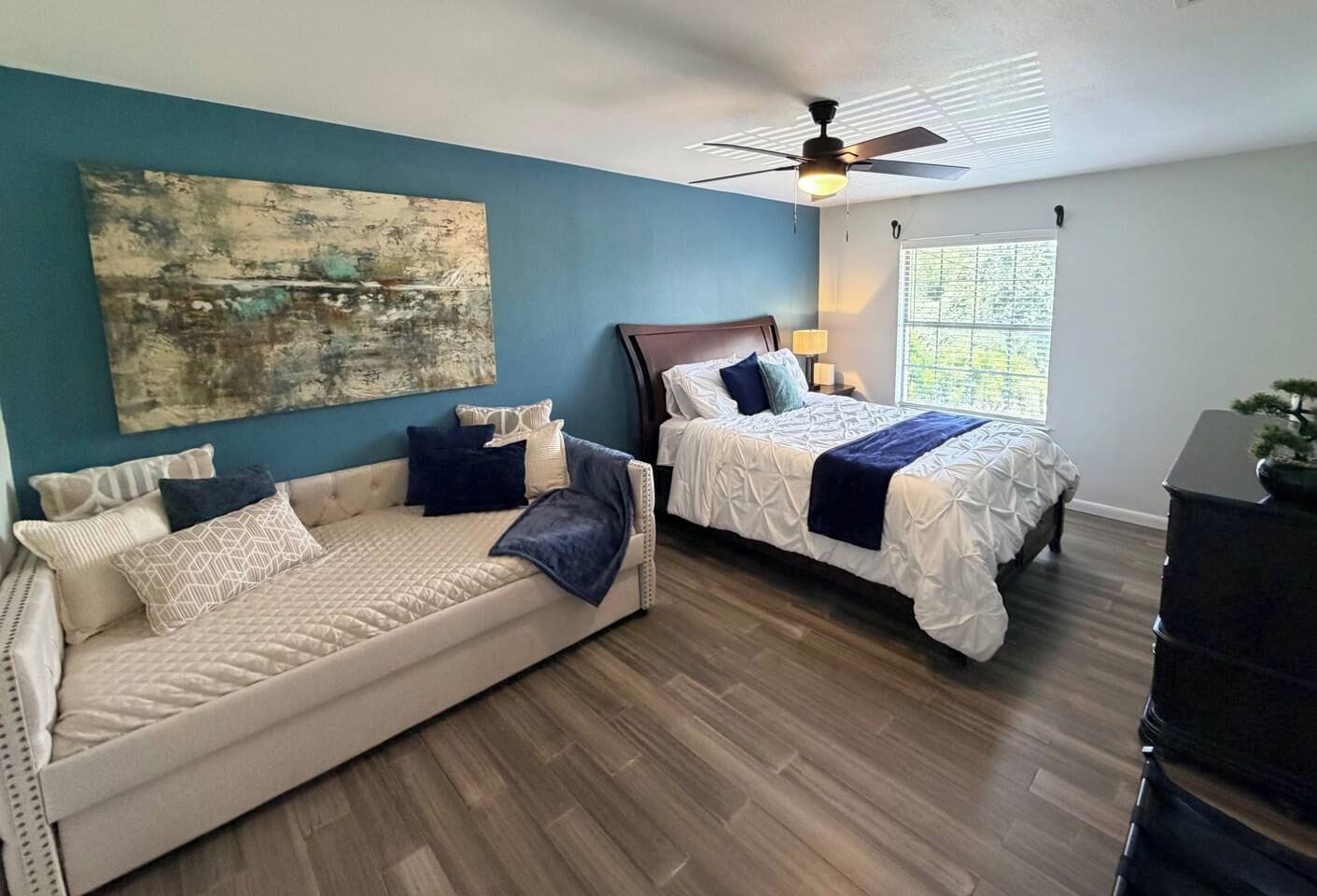A bedroom with a sofa, two beds, a ceiling fan, and a window with blinds.