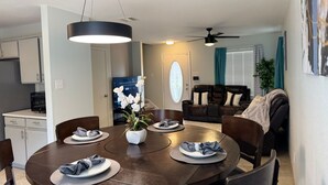 Ein Essbereich mit einem runden Tisch, Stühlen und einer Blumenvase. Es gibt eine Küchenzeile mit Schränken und einer Mikrowelle. Im Hintergrund ist ein Wohnzimmer mit einer Couch und einem Deckenventilator zu sehen.