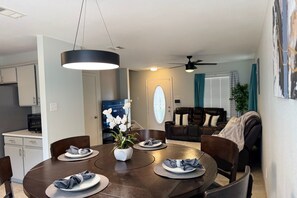 A dining area with a round table, chairs, and a vase of flowers. There is a kitchen area with cabinets and a microwave. A living room with a couch and a ceiling fan is visible in the background. - Austin Hotspot - Minutes to Airport, Downtown & F1 (Austin)
