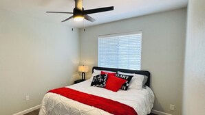 Ein Schlafzimmer mit Bett, Deckenventilator, Fenster mit Jalousien und Nachttisch mit Lampe.