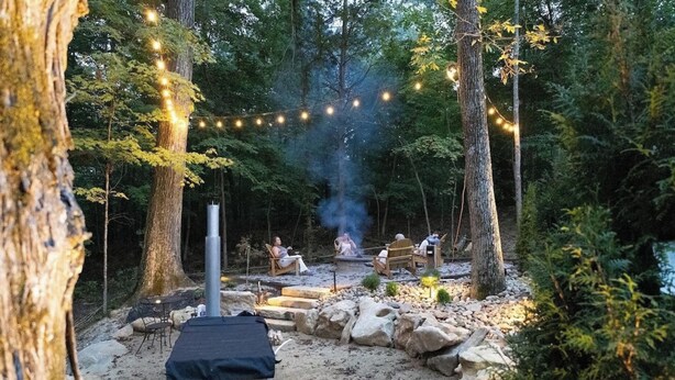 Un espace extérieur boisé, aménagé avec un foyer en pierre, des sièges et des guirlandes lumineuses.