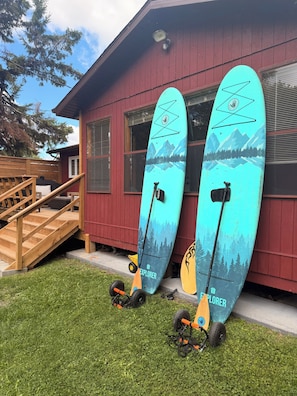 Deux planches de surf sont appuyées contre un bâtiment rouge, et un escalier en bois mène à une terrasse.