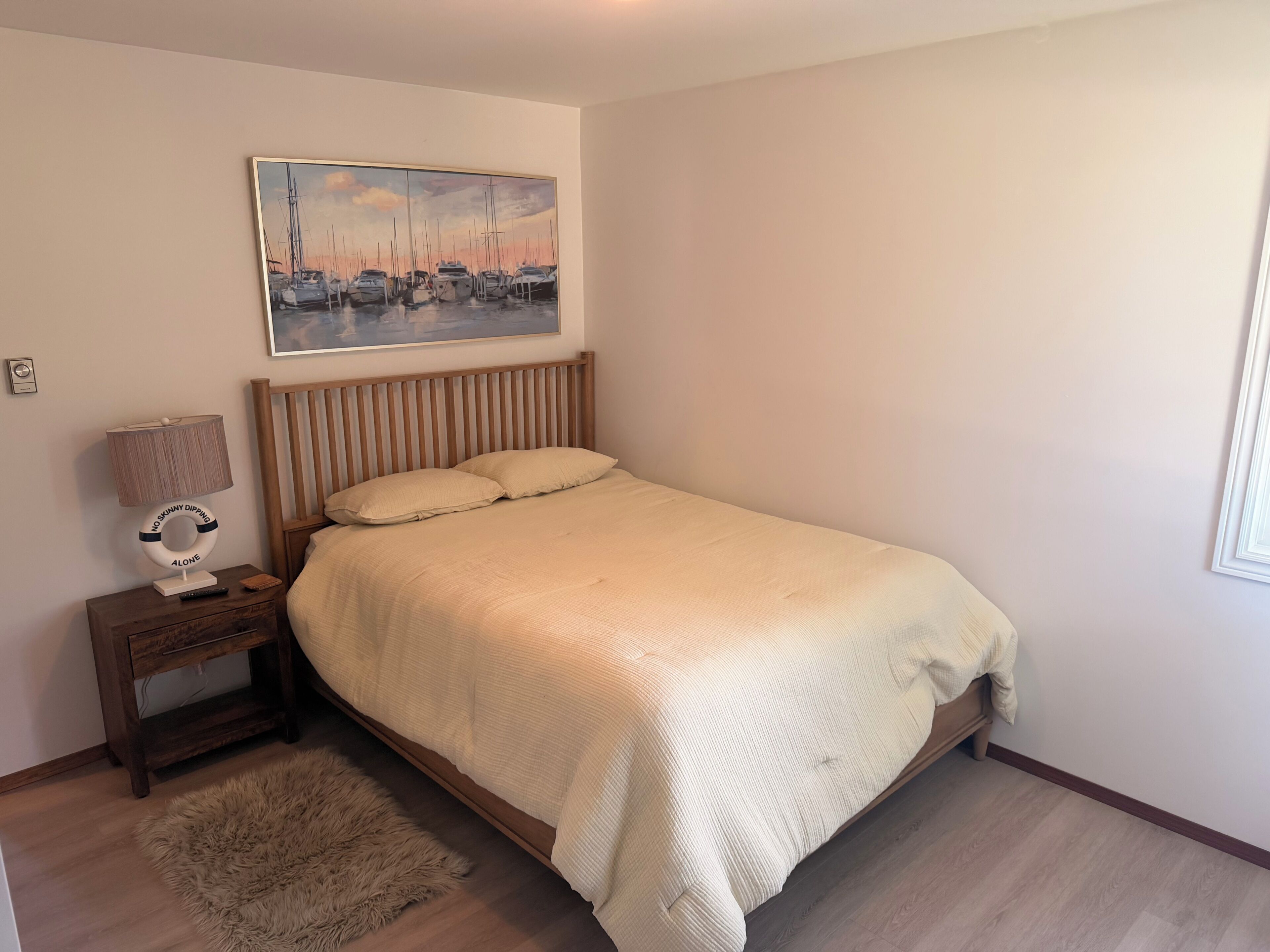 A bedroom with a bed, a nightstand, a lamp, a painting, and a fluffy rug.