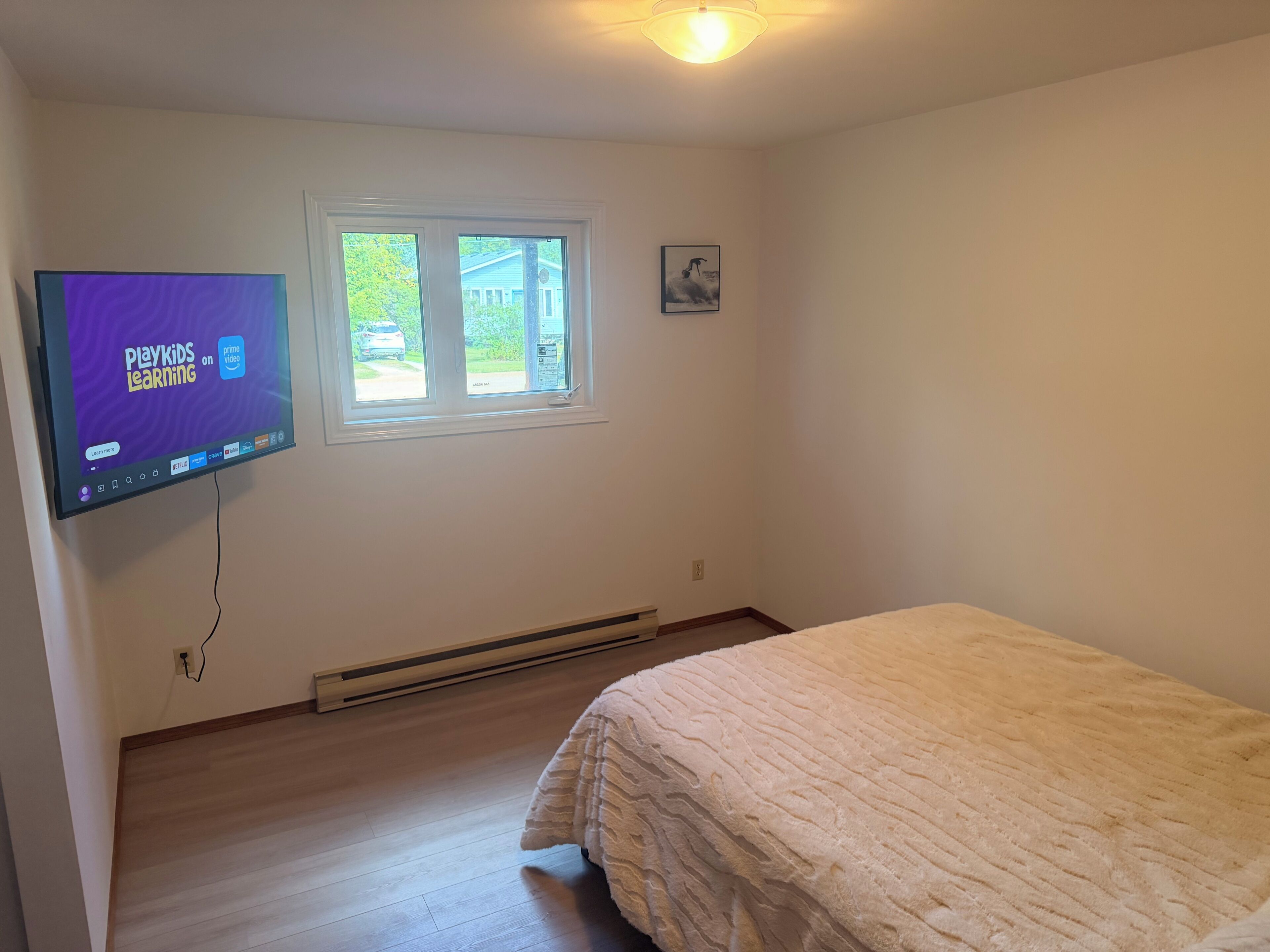 A bedroom with a bed, a wall-mounted TV displaying PlayKids Learning, a window, and a picture on the wall.