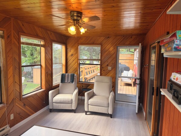 A room with wooden walls and ceiling, two beige armchairs, a ceiling fan, and a sliding glass door leading to an outdoor deck. - Cozy 3-bedroom cabin in adorable Gimli, Manitoba (Gimli)