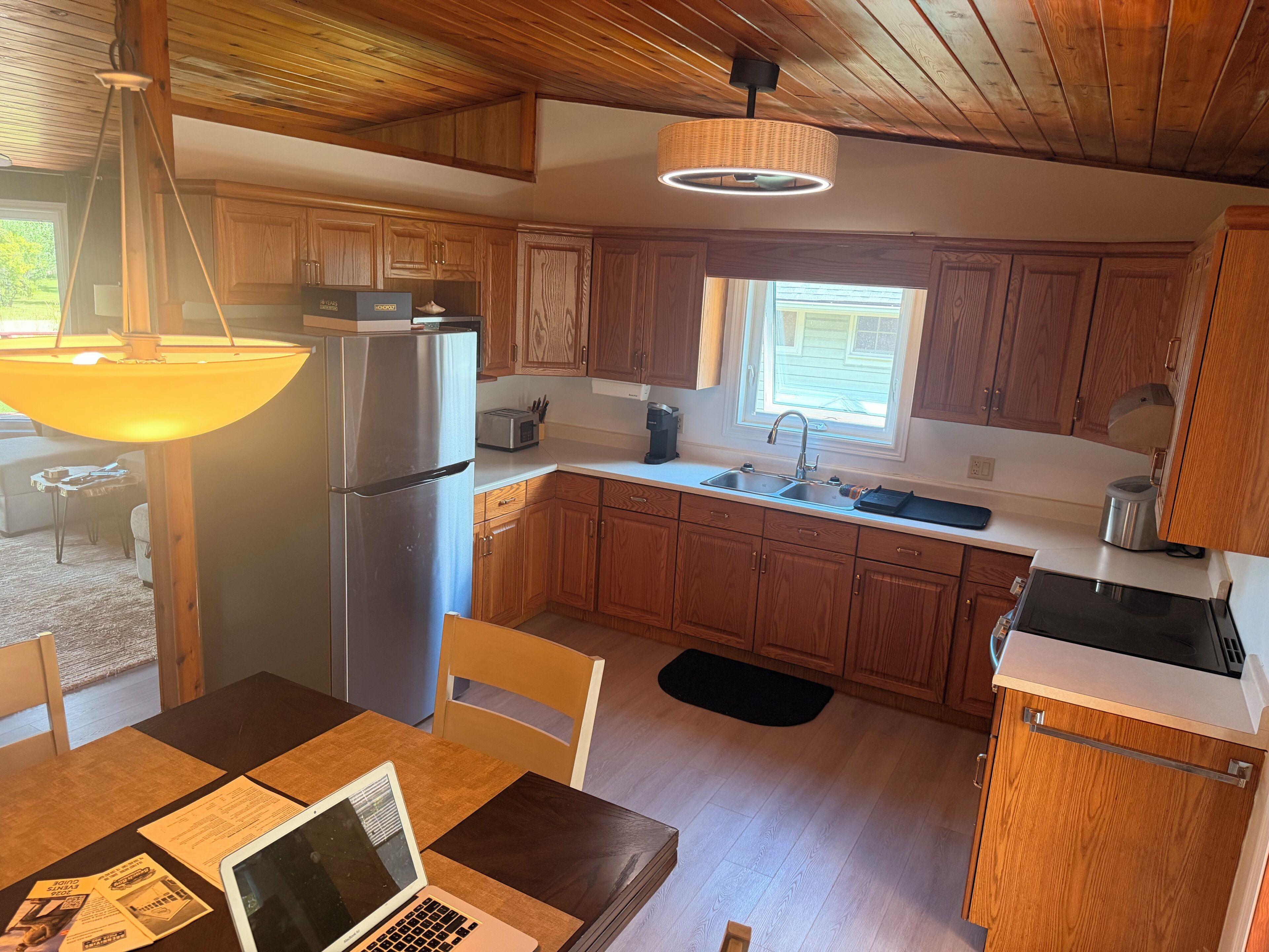 A kitchen with wooden cabinets, a stainless steel refrigerator, a sink, and a dining table with a laptop.