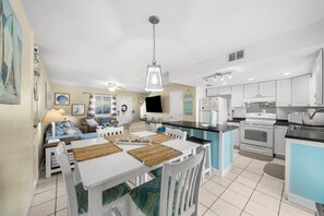A modern kitchen with white cabinets, a central island, and a dining area with white chairs and a table. - Captain’s Quarters @ Gulf Highlands Beach Resort (Panama City Beach)