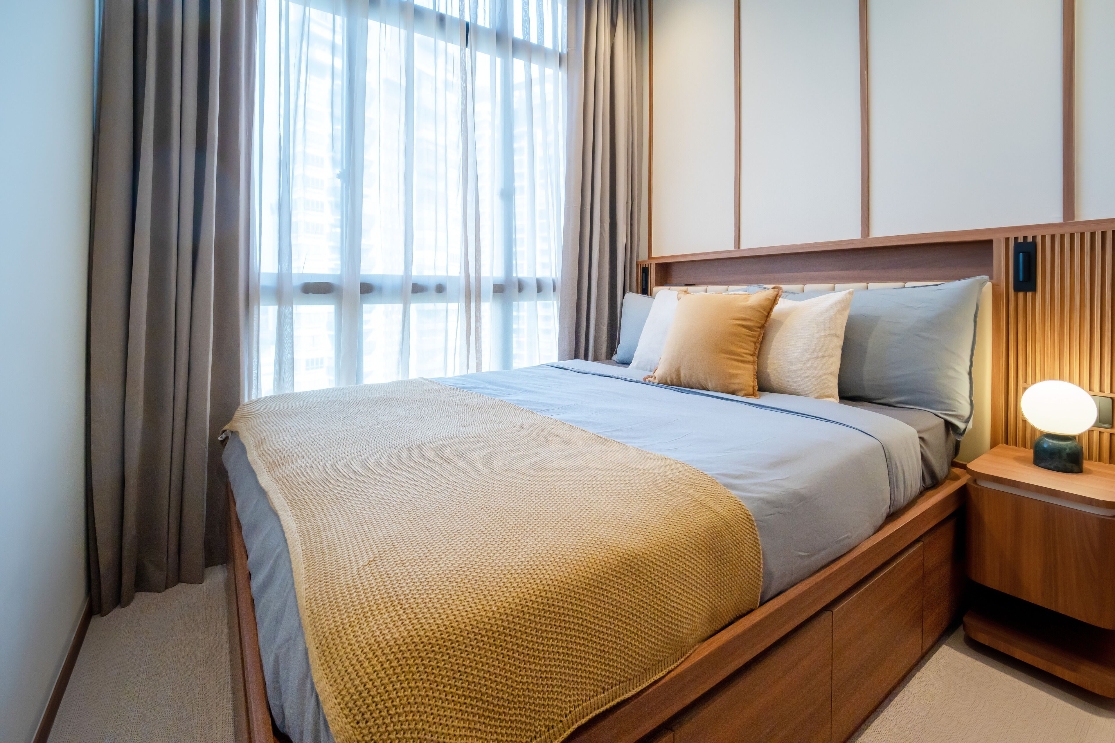 A neatly made bed with a wooden headboard, a bedside table with a lamp, and a view of the outside through sheer curtains.