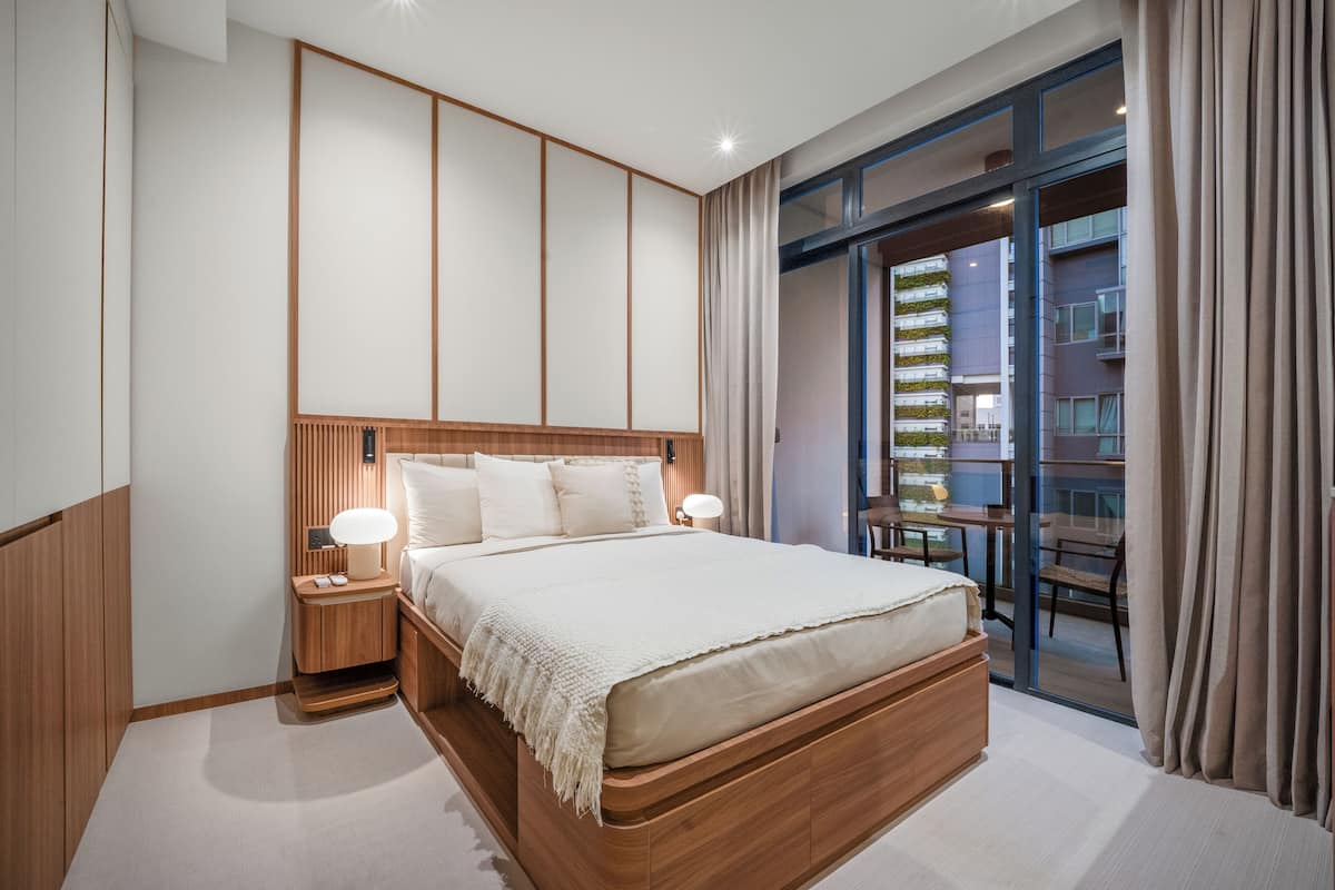 A modern hotel room with a large bed, wooden headboard, bedside lamps, and a view of a building through a glass door.