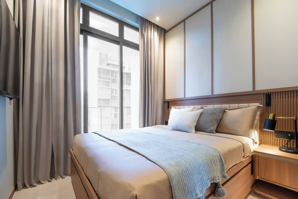 A modern hotel room with a large bed, a nightstand with a lamp, and a view of the city through large windows.