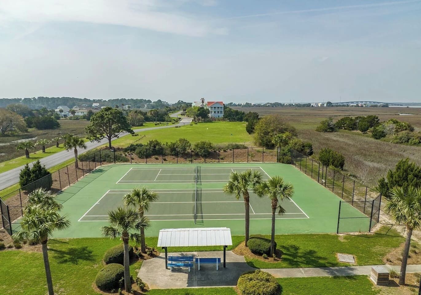 A tennis court with a surrounding fence, palm trees, and a building in the background.